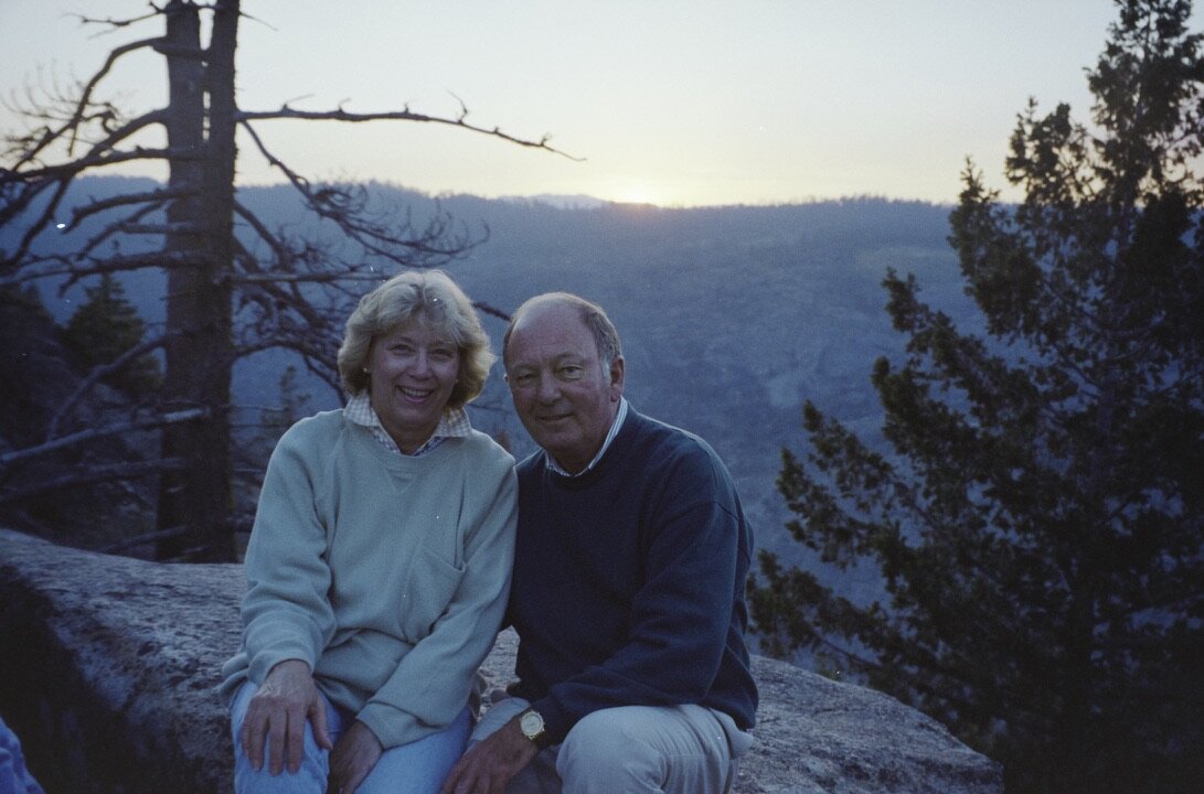 Jim and Jan sitting together on a granite ledge at sunset, a forested valley and distant ridgeline behind them near Camp Mather